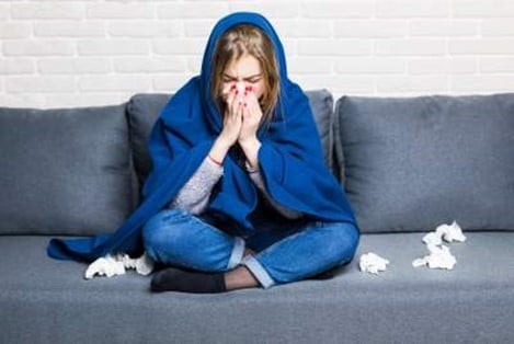 Female person sitting on a couch with a blue blanket over her shoulders. This image gives you the impression that this person is sick, and has a runny nose.